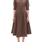 A woman wears the Simkhai Signature Jazz Dress—a brown cotton poplin midi with an A-line silhouette, collared neckline, elbow sleeves, and pleated skirt—paired with black open-toe sandals.