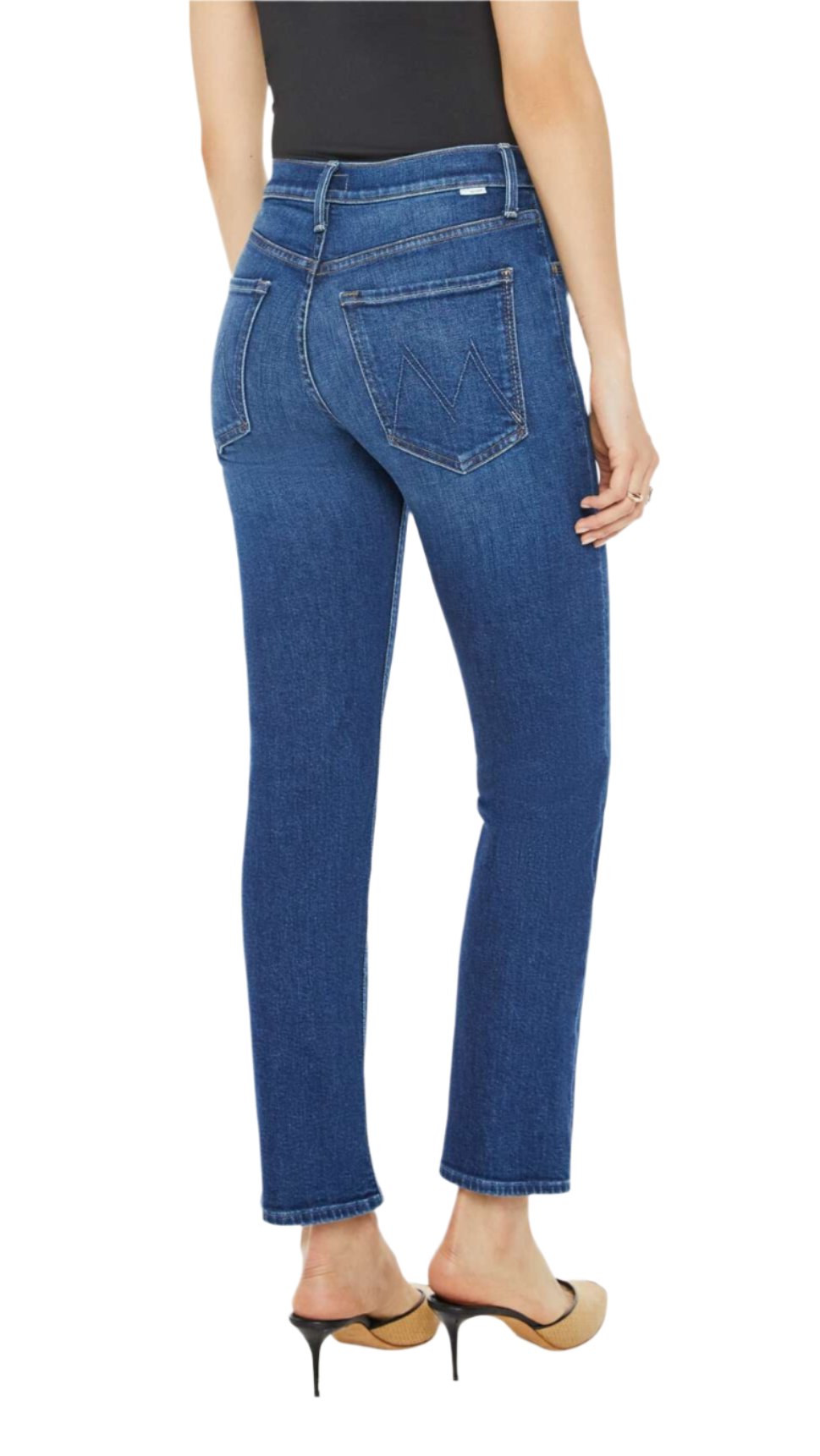 A person is standing sideways, wearing the Mid Rise Rider Flood by Mother—a black top with dark blue wash jeans featuring a straight leg cut. The stretch denim contours their silhouette beautifully, complemented by beige heeled sandals.