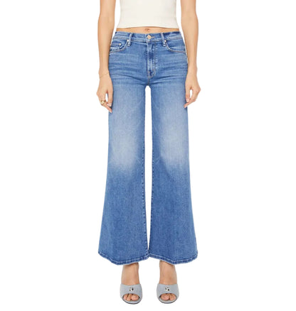 A person wears The Twister Skimp high-rise flared blue jeans by Mother, made from stretchy denim, with gray open-toe shoes, shown waist to feet against a white background.