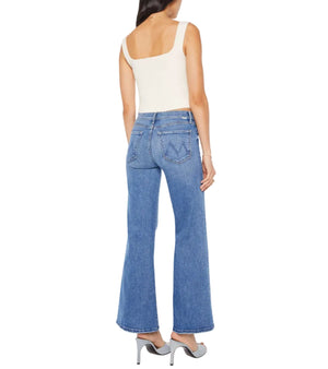 A woman stands with her back to the camera, wearing Mother’s The Twister Skimp blue high-rise flared wide leg jeans, a white tank top, and gray high heels against a white background.