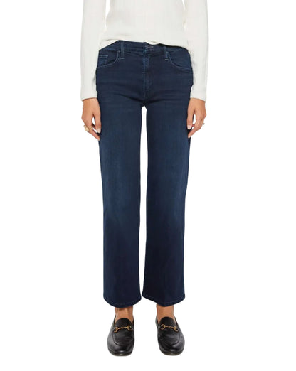 A person wearing Mother’s Mid Rise Rambler Zip Ankle jeans in dark blue stretch denim, paired with a white long-sleeve top and black loafers, standing against a plain background.
