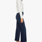 A woman stands in profile against a white background, wearing a long-sleeve white top, black loafers, and the Mother Mid Rise Rambler Zip Ankle jeans—dark blue, mid-rise wide straight leg jeans crafted from stretch denim.