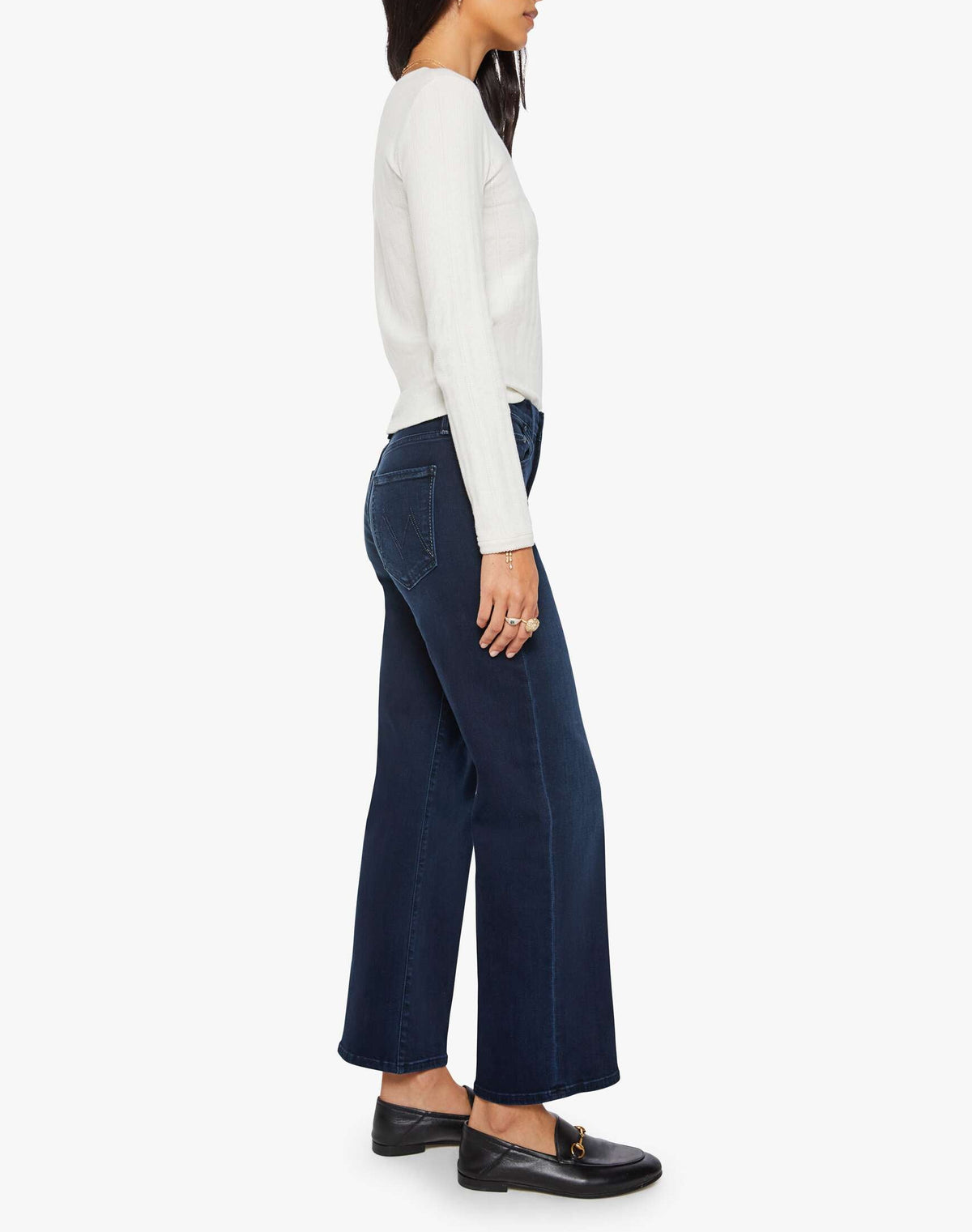 A woman stands in profile against a white background, wearing a long-sleeve white top, black loafers, and the Mother Mid Rise Rambler Zip Ankle jeans—dark blue, mid-rise wide straight leg jeans crafted from stretch denim.