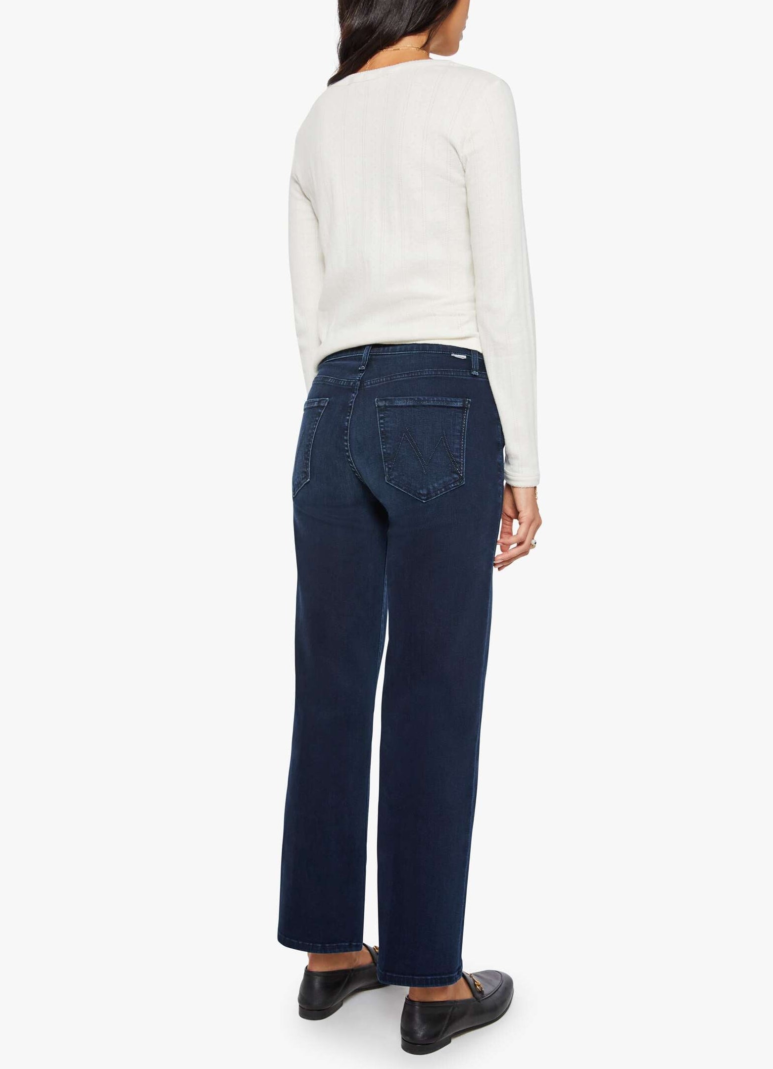 A person is wearing a long-sleeve white top, black shoes, and the Mother Mid Rise Rambler Zip Ankle jeans—dark blue stretch denim with a wide straight leg cut and mid-rise fit—standing with their back to the camera against a plain white background.