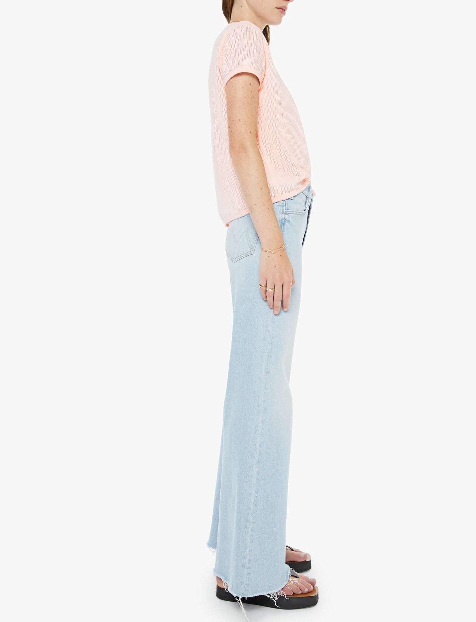 A person standing in profile wears a light pink short-sleeved shirt, The Hustler Roller Skimp Fray jeans by Mother in a light blue high-rise wide-leg style, and black sandals against a plain white background.