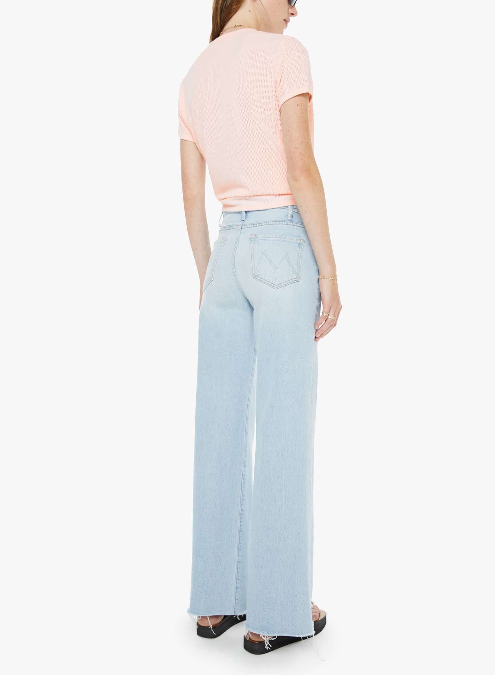 Facing away from the camera against a plain white background, the person is wearing The Hustler Roller Skimp Fray jeans by Mother in a light blue wash, paired with a light pink short-sleeve top and black sandals.