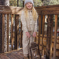 A young girl wearing the Rylee & Cru Girls’ Denim Chore Coat, styled over leopard-print pajamas and a gray beanie, stands on a wooden deck holding a vintage lantern, with trees and a wooden fence in the background.