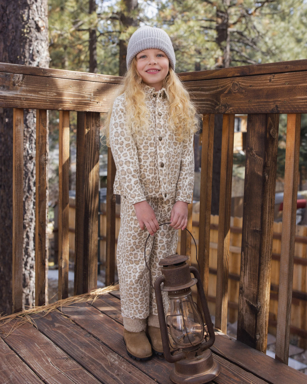 A young girl wearing the Rylee & Cru Girls’ Denim Chore Coat, styled over leopard-print pajamas and a gray beanie, stands on a wooden deck holding a vintage lantern, with trees and a wooden fence in the background.