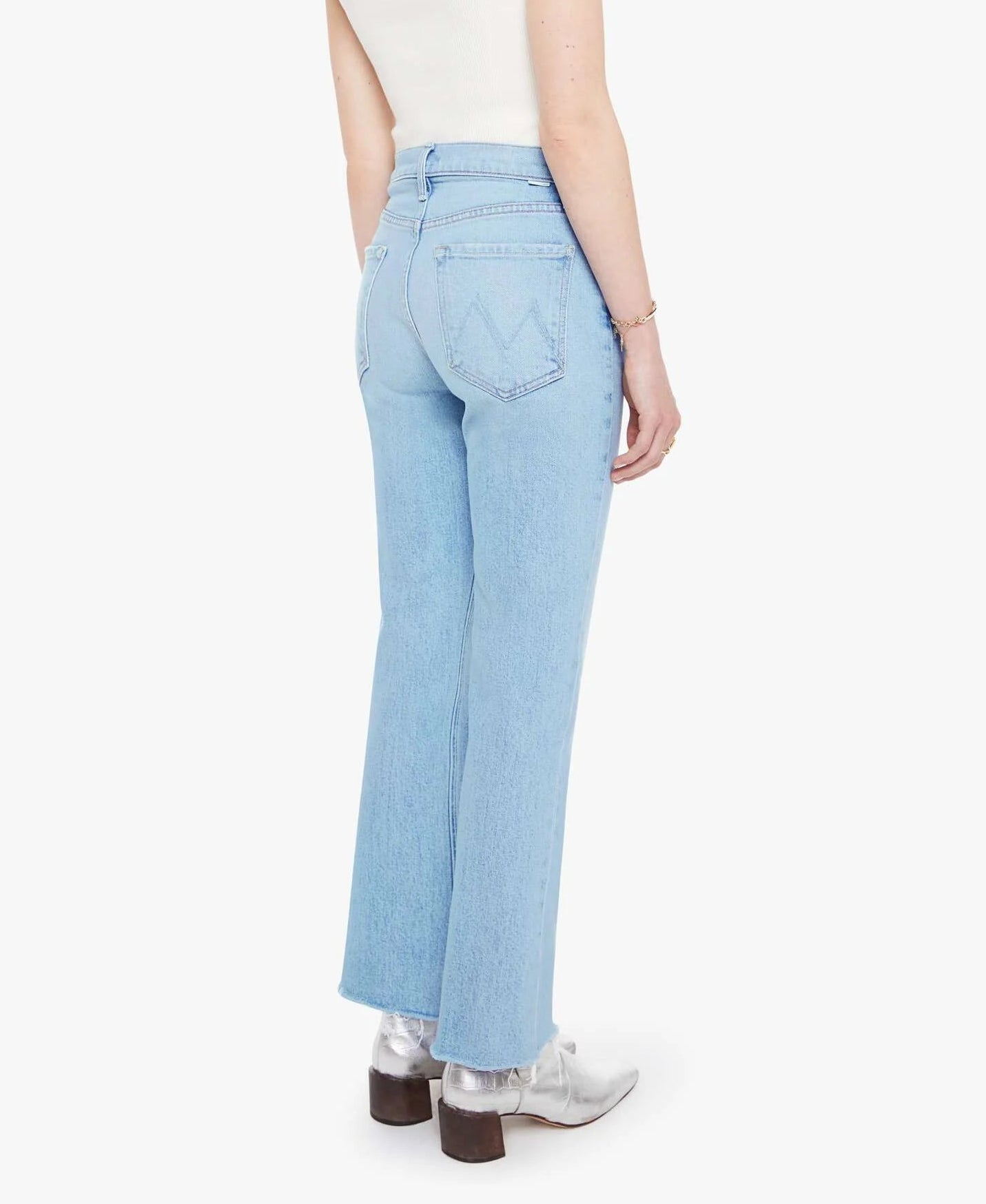 A person wearing Mother’s The Tripper Flood Fray light blue high-rise flare jeans, a white sleeveless top, and silver ankle boots stands with their back to the camera against a plain white background.