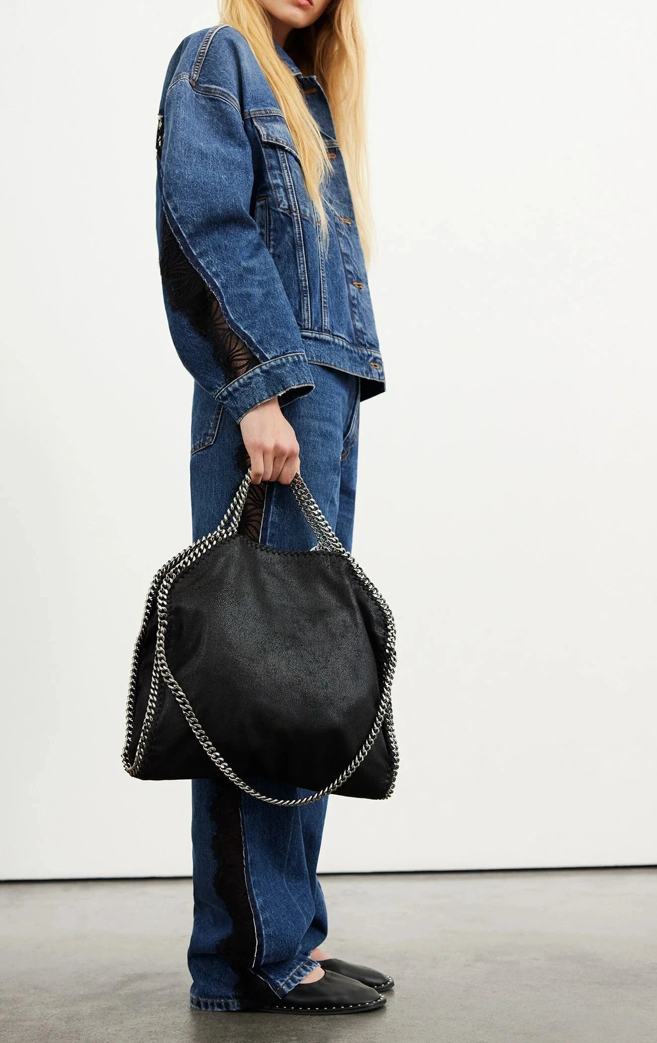 A person in a blue denim jacket and jeans holds a Stella McCartney Falabella Fold Over Tote with chain handles, made from recycled plastic bottles; only their lower face and body are visible.