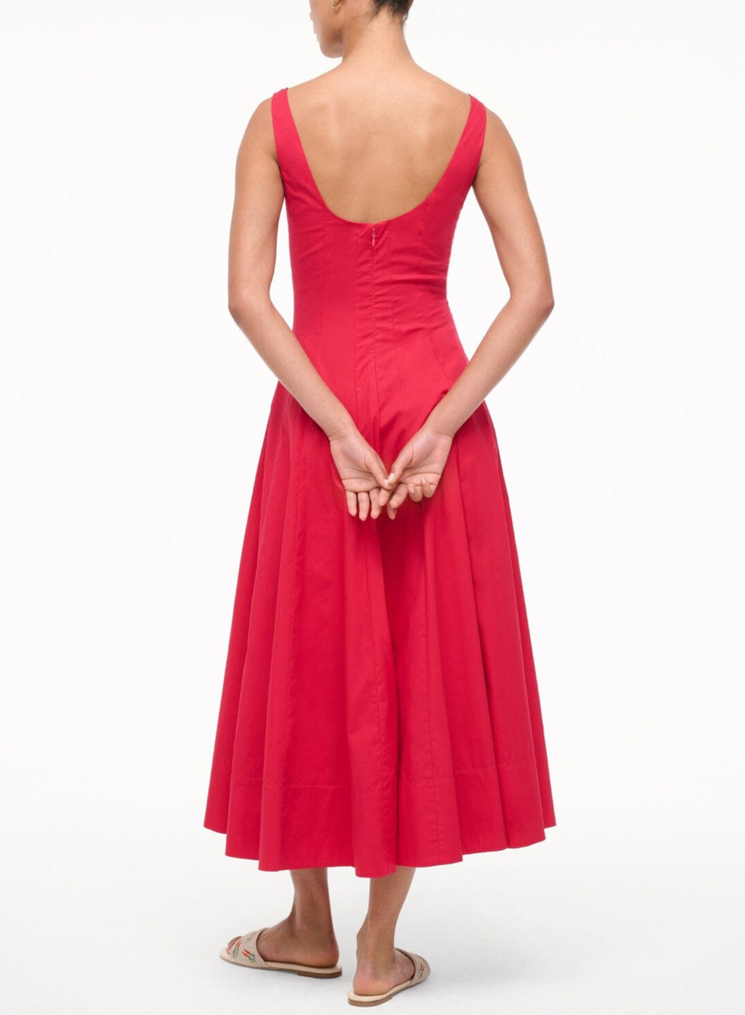 A woman, her back to the camera, wears the Staud Wells Dress—a bright red, ankle-length cotton poplin style with a flattering corset-inspired panel. Her hands are clasped behind her back and she's wearing flat sandals.