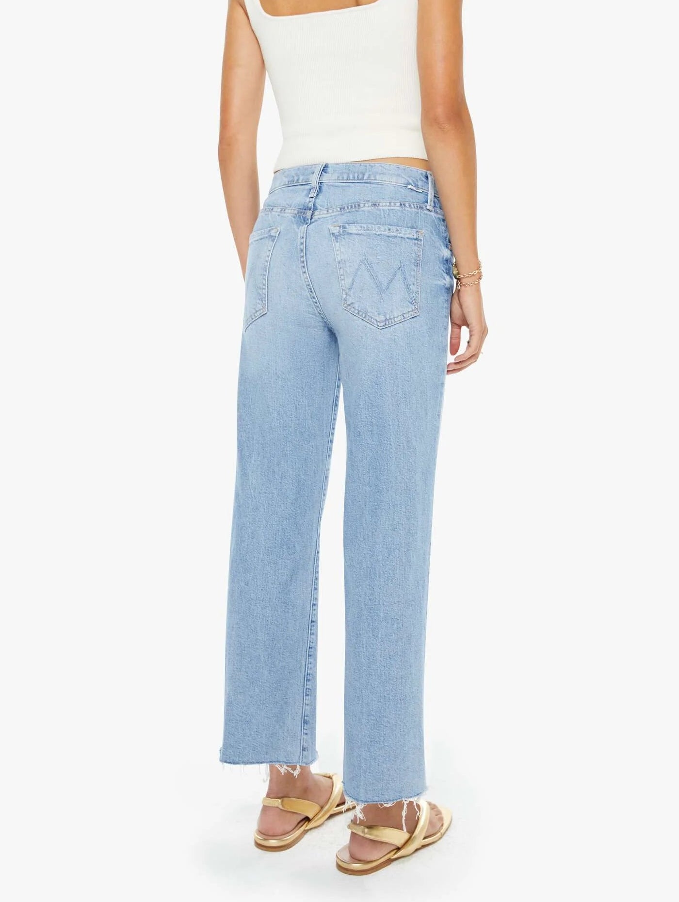 A woman is shown from the back wearing Mother’s Mid Rise Rambler Zip Ankle Fray jeans in light blue with a wide straight leg and frayed hem, paired with a white sleeveless top and sandals against a plain background.