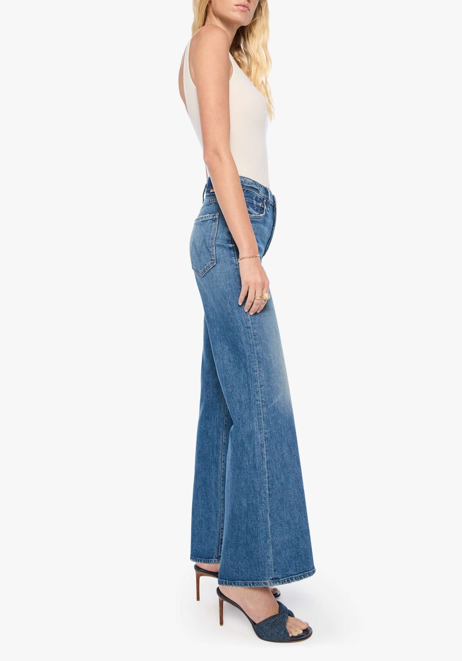 A woman stands sideways against a plain white background wearing heeled sandals, a white sleeveless top, and the Tomcat Roller Sneak high-waisted wide-leg jeans by Mother in a vintage-inspired blue wash.