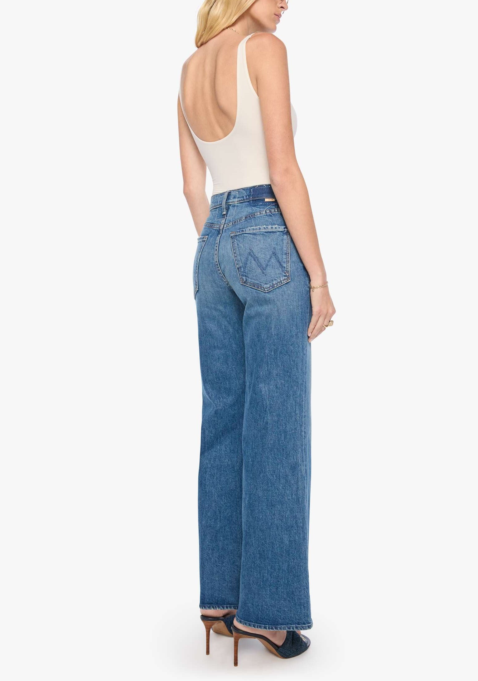 A woman stands in profile against a white background, showcasing the Mother Tomcat Roller Sneak jeans—a high-waisted, wide-leg silhouette crafted from vintage-inspired SUPERIOR denim—paired with a white sleeveless bodysuit and high heels, her back partially visible.