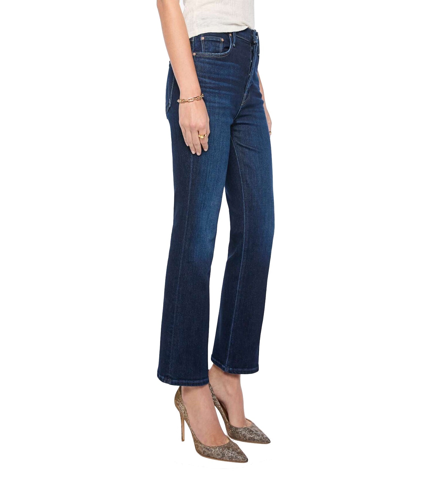 Wearing the Mother Tripper Flood dark blue high-rise flare bootcut jeans, styled with a white top, gold bracelet, and pointed glittery heels, this person stands against a white background.