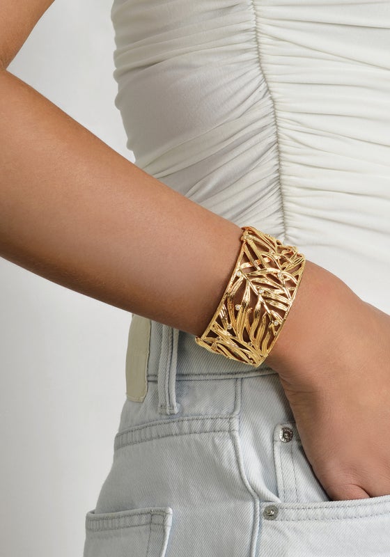 A person wears a white ruched top and light denim jeans, accessorized with the Anabel Aram Palm Leaves Cuff—an 18k gold-dipped bracelet styled like palm fronds.