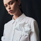A woman with straight brown hair models the Draped Rose Button Down Shirt by Lela Rose, a white cotton poplin shirt with a large fabric bow on the chest, standing against a dark background.