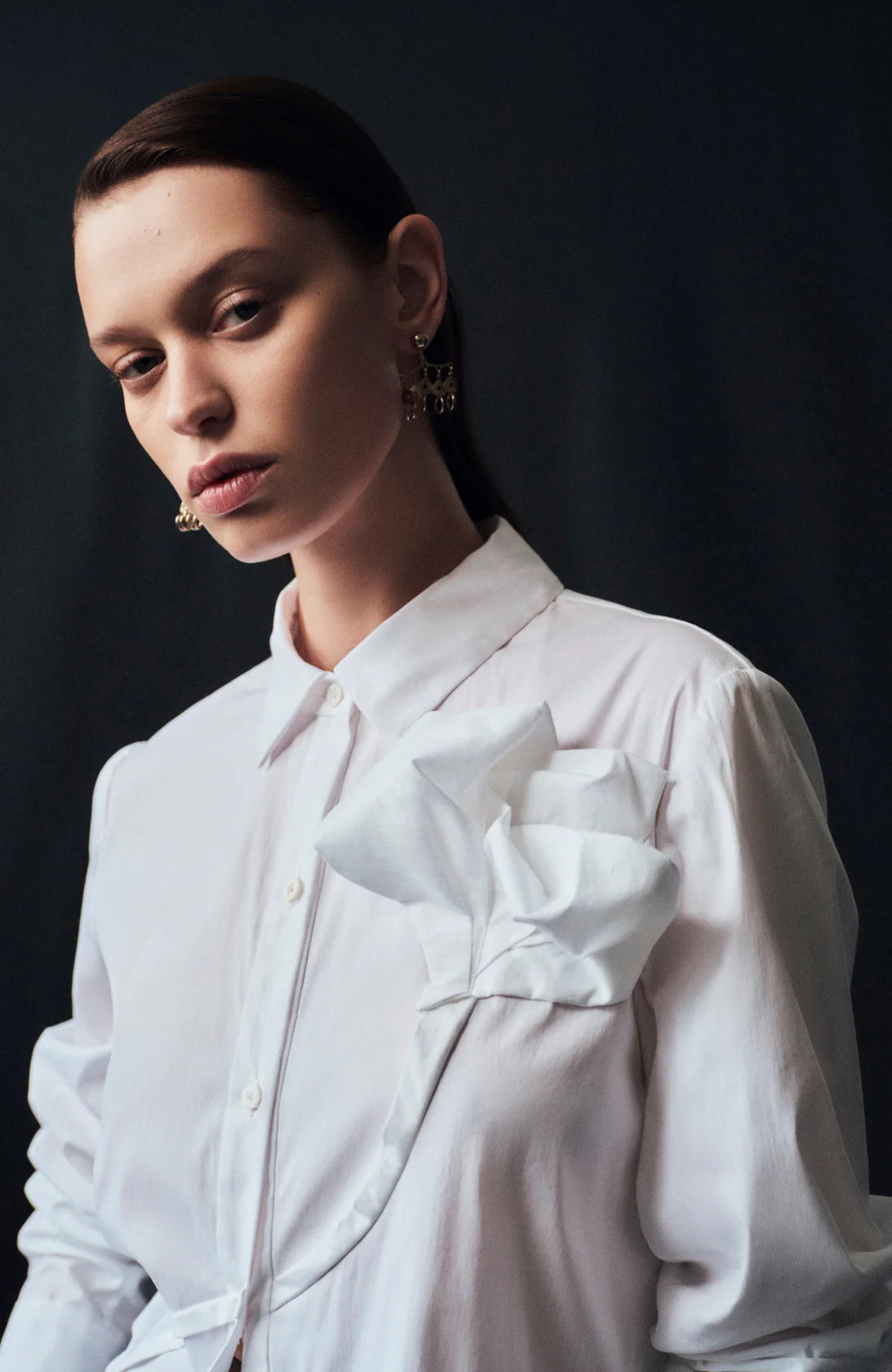 A woman with straight brown hair models the Draped Rose Button Down Shirt by Lela Rose, a white cotton poplin shirt with a large fabric bow on the chest, standing against a dark background.