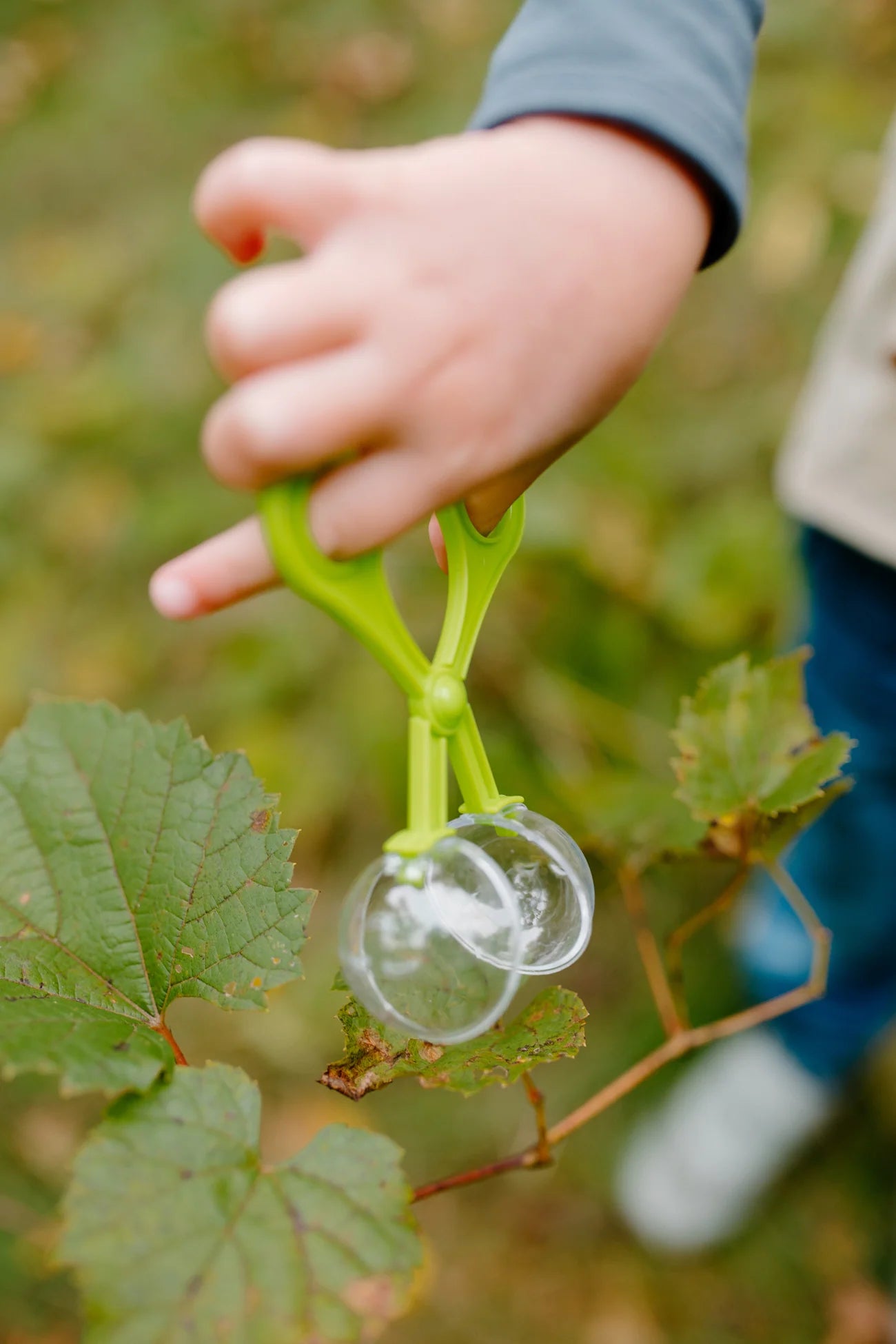 A child’s hand, dressed in the Great Pretenders Forest Guardian Set with Accessories, uses green plastic tweezers to pick up a transparent object near green leaves outdoors.