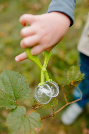 A child’s hand, dressed in the Great Pretenders Forest Guardian Set with Accessories, uses green plastic tweezers to pick up a transparent object near green leaves outdoors.