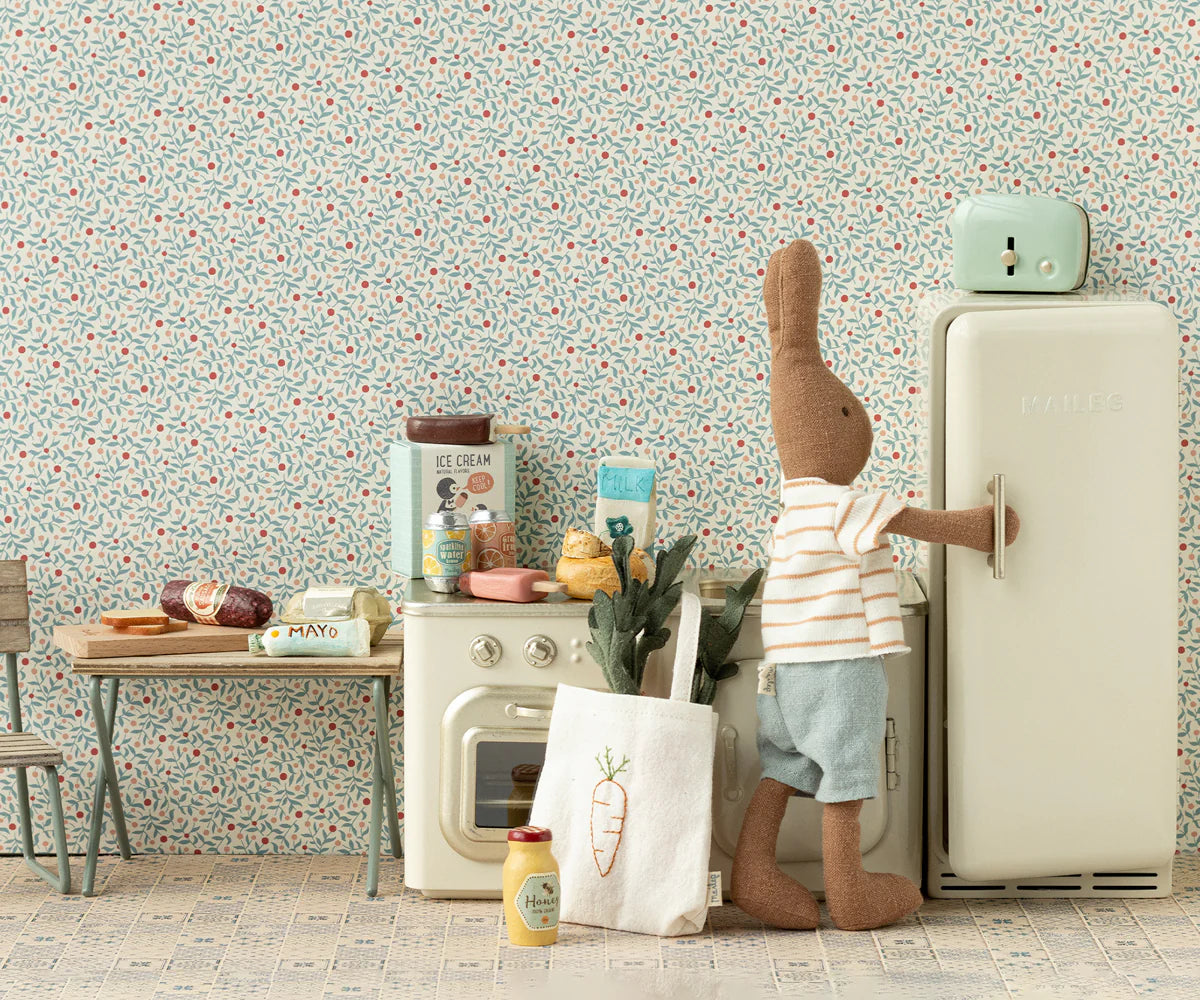 A Maileg stuffed bunny in a striped shirt and shorts stands by a metal toy fridge in the Maileg Miniature Kitchen, surrounded by play food, kitchen items, and fresh buns.