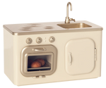 The Maileg Miniature Kitchen is a compact set featuring a beige and gold stove, oven with baked buns, sink, and cupboard.