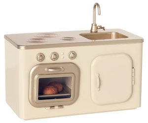 The Maileg Miniature Kitchen is a compact set featuring a beige and gold stove, oven with baked buns, sink, and cupboard.