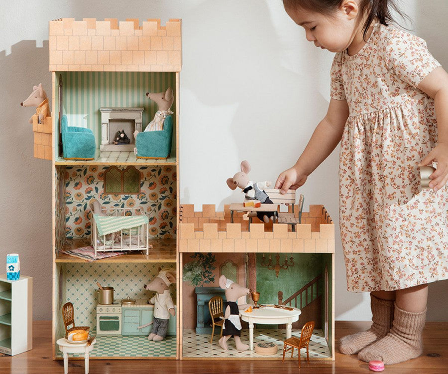 A child plays with a miniature dollhouse featuring small toy mice in different rooms, including a living room with the Maileg Fireplace in Off-White, nursery, and dining area. The vintage painted look of this castle collection shines as the child arranges a mouse on the balcony.