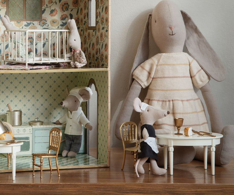 A detailed dollhouse with three rooms showcases stuffed toy mice engaged in various activities, including a Maileg Dining Table set for a cheese feast. Beside the dollhouse sits a large stuffed rabbit, adding to the charming scene.