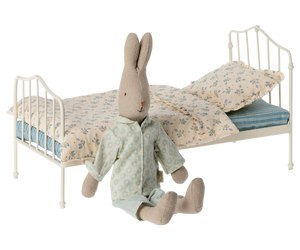 A Maileg Miniature Bed, Mini - Blue features floral bedding and a striped mattress, with a stuffed rabbit in pajamas sitting in front, creating a charming vintage look.