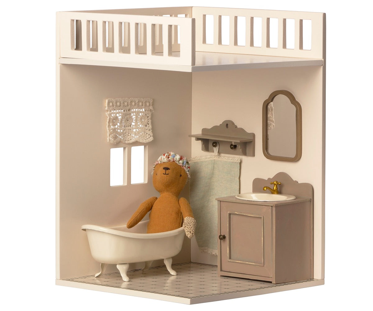 The Maileg House of Miniature Bathroom showcases a delightful scene with a charming teddy bear lounging in a classic clawfoot bathtub, complemented by elegant dollhouse furniture such as a sink with mirror, a side towel, and a delicate lace curtain framing the window.