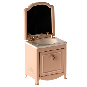 The Maileg Sink Dresser, Mouse - Powder is a small pink vanity with a mirror, gold faucet, and cabinet door below the bathroom sink.