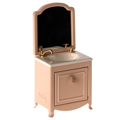 The Maileg Sink Dresser, Mouse - Powder is a small pink vanity with a mirror, gold faucet, and cabinet door below the bathroom sink.