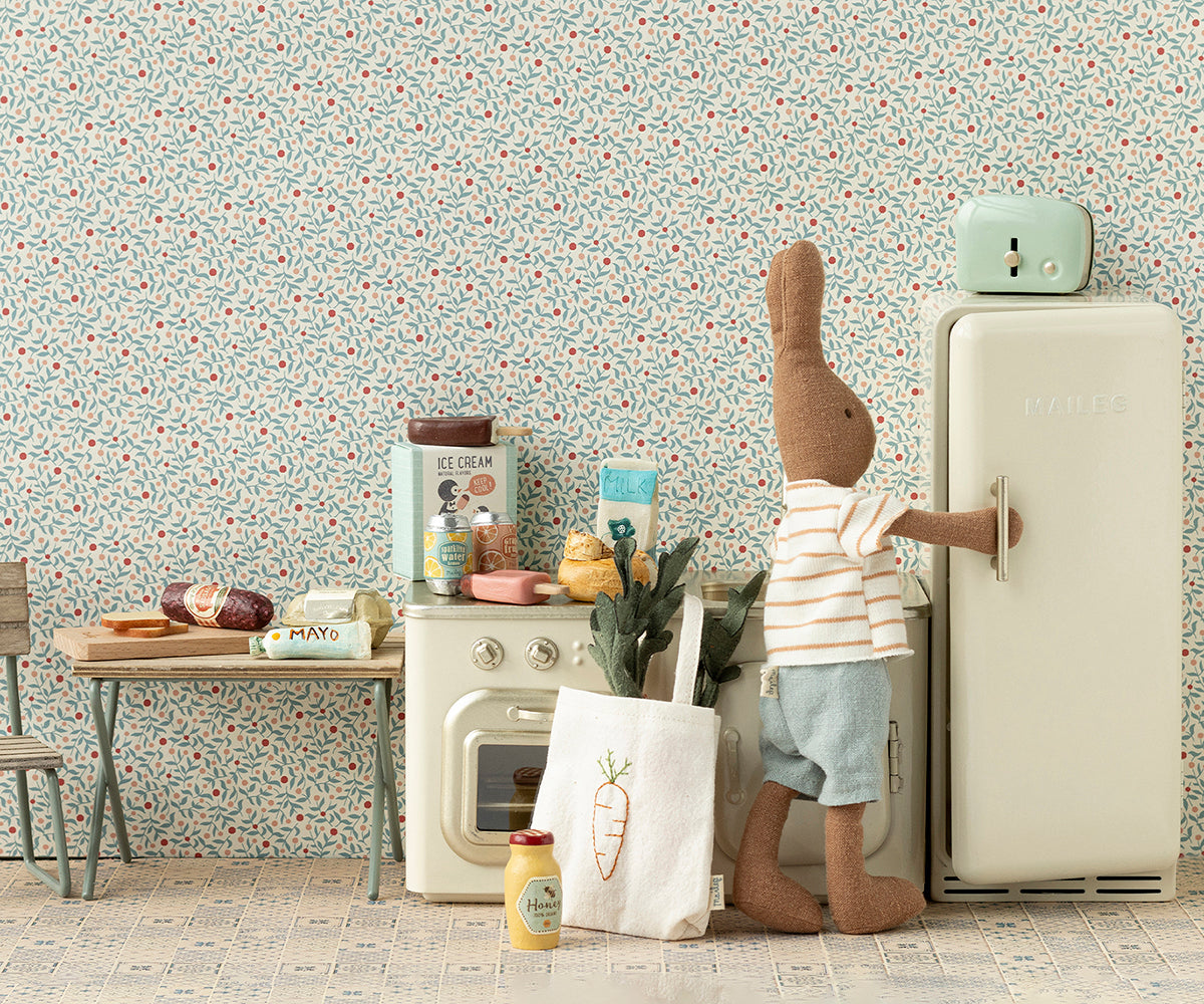 A Maileg stuffed rabbit in a striped shirt and shorts opens the Maileg Miniature Fridge in a vintage-style miniature kitchen filled with various food items and appliances.