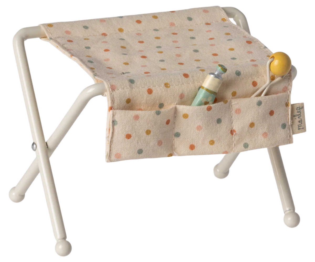 The Maileg Nursery Table, Baby Mouse - Off White, features a beige polka-dot seat with pockets to hold accessories like a baby bottle, yellow pacifier, and tiny rattle—ideal for a cozy baby mice nursery setup.