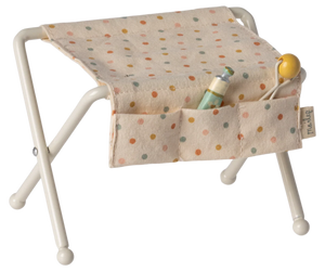 The Maileg Nursery Table, Baby Mouse - Off White, features a beige polka-dot seat with pockets to hold accessories like a baby bottle, yellow pacifier, and tiny rattle—ideal for a cozy baby mice nursery setup.