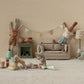 Stuffed rabbits in a living room set: one on a couch, another hanging bunting at the table. Others wrap gifts and tinker with decorations. Cozy decor includes a lamp, plants, and a mini cake atop Maileg's Cakes and Cakestand, Mini, enhancing the whimsical ambiance of this teddy family gathering.