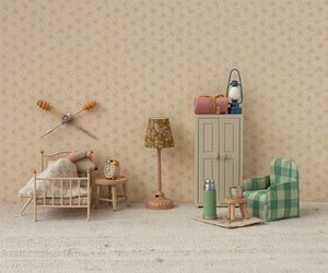 A miniature bedroom scene featuring a bed, pillows, alarm clock, Maileg Vintage Floor Lamp, Mouse - Dark Powder casting a soft light, wardrobe, stacked bags, green armchair, small table, thermos, cup, and decorative wall hooks.