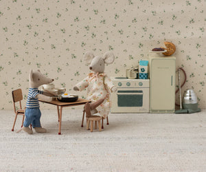Two toy mice, one sitting at a Maileg Table & Chair Set, Mouse - Dark powder, and one standing, are in a small vintage-style toy kitchen surrounded by miniature appliances and furniture.