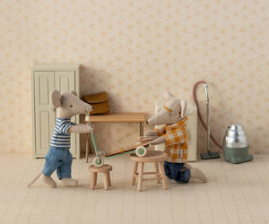 Two magnetic mice in casual clothes assemble the Maileg Kick Board, Mouse - Mint—a green scooter with wooden details—in a miniature room featuring stools, a vacuum cleaner, and a cabinet.
