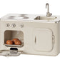 The Maileg Kitchen - Miniature is a charming metal kitchen set that includes a stovetop, sink, and an oven with an open door displaying a tray of three round pastries inside. Designed in perfect dollhouse scale, its metallic countertop and knobs add to the authentic detail.