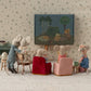 Maileg friends gather in their whimsical Mouse Hole Farmhouse, with stuffed mice toys sitting on assembled Folding Chair, Mouse - Rose. They watch a small fabric TV showing a camping scene, surrounded by cozy furniture and decorative wallpaper.