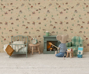 A Maileg Folding Chair, Mouse - Green Check sits in a miniature Mouse hole Farmhouse living room, surrounded by a bed, fireplace, table, food, and a patterned wall featuring animals and desert plants.