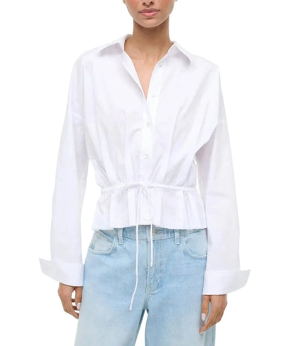 A person wears the Staud Primrose Shirt, a white poplin button-down with a waisted drawstring, paired with light blue jeans, shown from mid-thigh up.