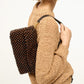 A person in a beige knit sweater carries the Staud Timmy Convertible Shoulder Bag—rectangular, dark brown beaded design with a black strap and interior zip pocket—over their shoulder.
