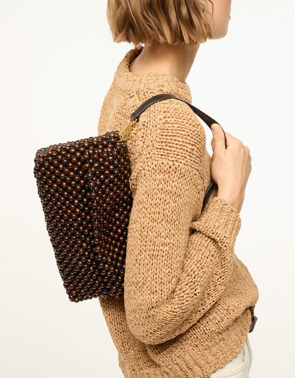 A person in a beige knit sweater carries the Staud Timmy Convertible Shoulder Bag—rectangular, dark brown beaded design with a black strap and interior zip pocket—over their shoulder.