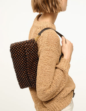 A person in a beige knit sweater carries the Staud Timmy Convertible Shoulder Bag—rectangular, dark brown beaded design with a black strap and interior zip pocket—over their shoulder.