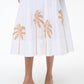 A person wearing the Staud Wells Palm Embroidery Dress, a white cotton poplin midi dress with brown palm tree embroidery at the hem and a fitted waist, paired with brown heeled sandals.