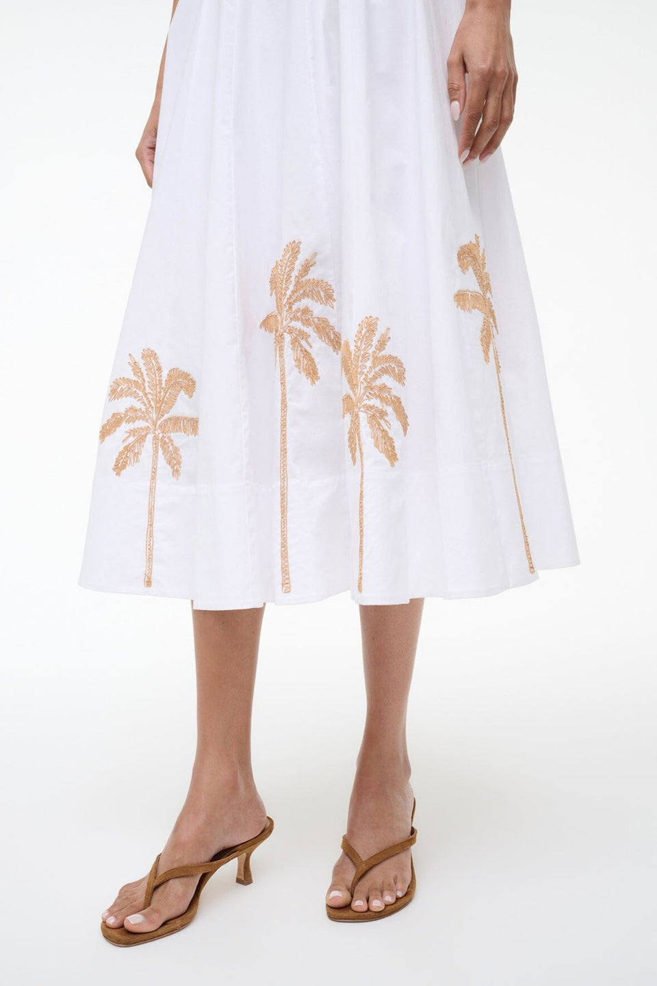 A person wearing the Staud Wells Palm Embroidery Dress, a white cotton poplin midi dress with brown palm tree embroidery at the hem and a fitted waist, paired with brown heeled sandals.