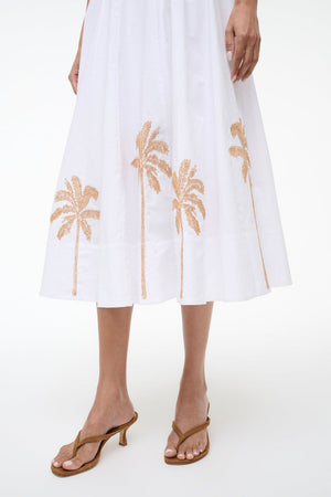 A person wearing the Staud Wells Palm Embroidery Dress, a white cotton poplin midi dress with brown palm tree embroidery at the hem and a fitted waist, paired with brown heeled sandals.